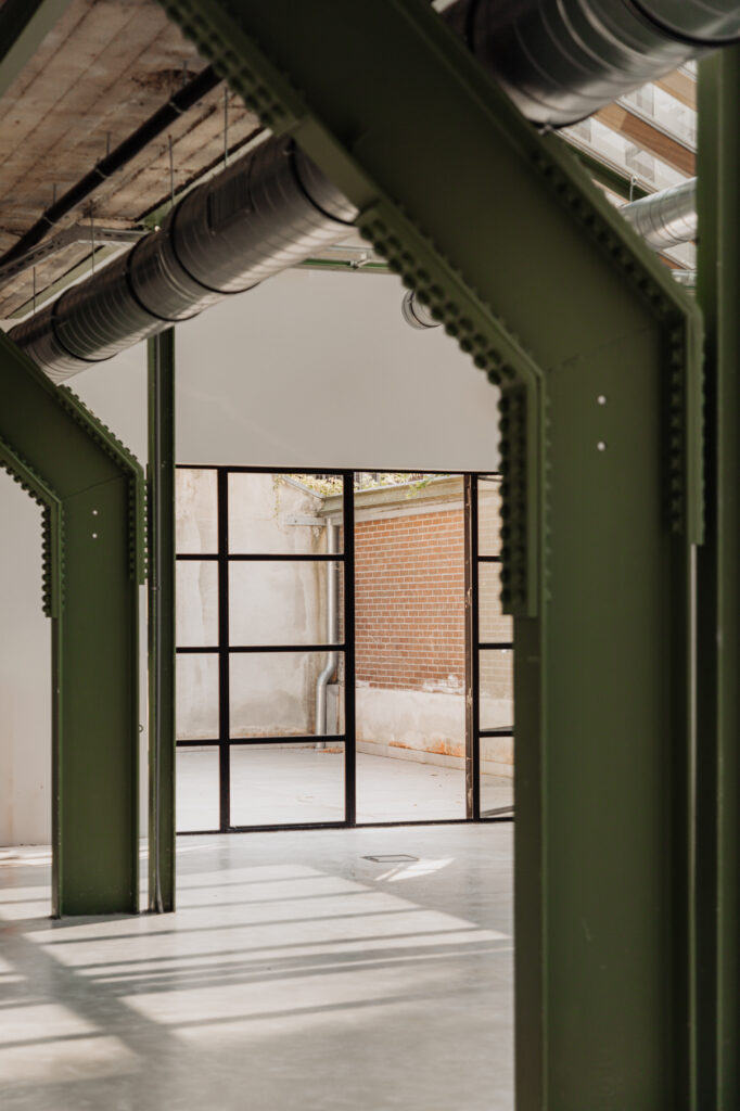 Industriële binnenruimte met groene stalen balken, zicht op glazen deuren en een binnentuin aan de Plantage Middenlaan.